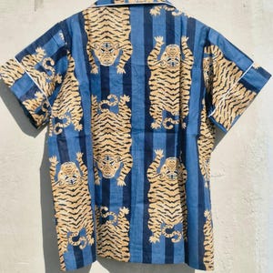 May include: Short-sleeved shirt with a blue and white striped pattern. The shirt features a repeating design of stylized tigers in shades of tan and brown. White trim accents the collar and sleeves. The shirt is hanging on a wooden hanger.