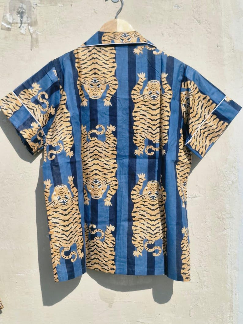 May include: Short-sleeved shirt with a blue and white striped pattern. The shirt features a repeating design of stylized tigers in shades of tan and brown. White trim accents the collar and sleeves. The shirt is hanging on a wooden hanger.