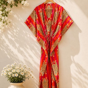 Puede incluir: Un vestido caftán largo hasta el suelo con un vibrante estampado de tigre en tonos naranja, rosa y negro. La prenda presenta un escote en V profundo, mangas anchas estilo kimono y un cordón ajustable en la cintura que crea una silueta fruncida.
