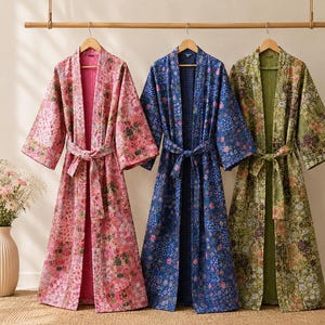 May include: Three long-sleeved robes with matching tie belts, each featuring a distinct floral pattern in shades of pink, blue, and green. The robes have an open front design, wide sleeves, and a quilted texture, hanging side by side on wooden hangers.