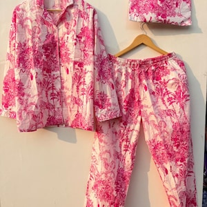 May include: A pink and white pajama set with a matching sleep mask. The set includes a button-down shirt, pants, and a sleep mask, all featuring a pink floral and bird pattern. The pajamas are hanging on wooden hangers.
