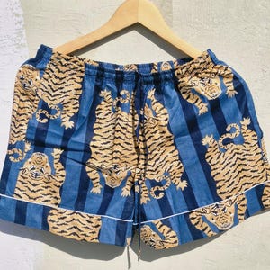 May include: Blue and tan tiger print shorts with a drawstring waist and white trim. The shorts are hanging on a wooden hanger. The fabric features a repeating pattern of tigers and blue vertical stripes.