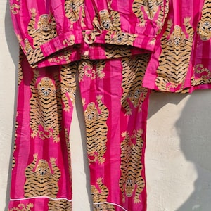 May include: A pink pajama set with a tiger print. The set includes a top and pants, both featuring a repeating pattern of tigers in shades of brown and black against a pink striped background. White trim accents the edges.