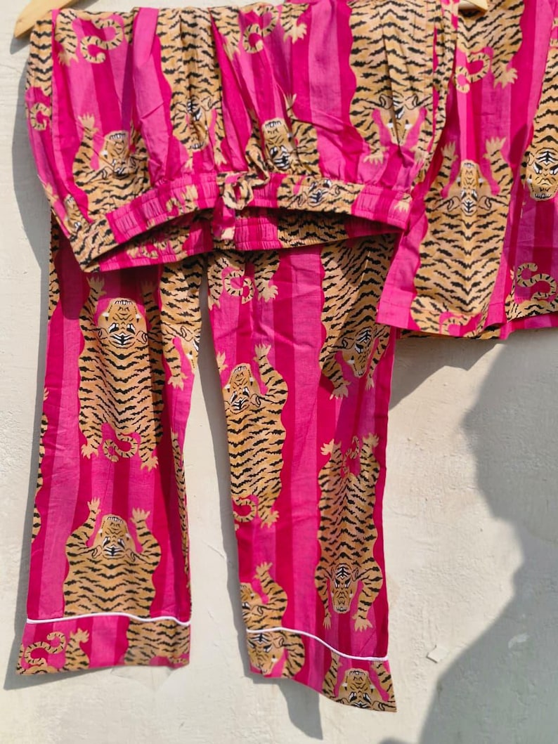May include: A pink pajama set with a tiger print. The set includes a top and pants, both featuring a repeating pattern of tigers in shades of brown and black against a pink striped background. White trim accents the edges.
