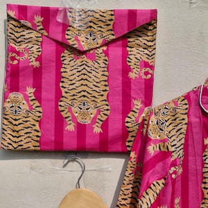 May include: Pink and brown tiger print pajama set. The set includes a button-down shirt and matching pants, with a repeating tiger pattern on a pink striped background. The set is displayed with a matching envelope.