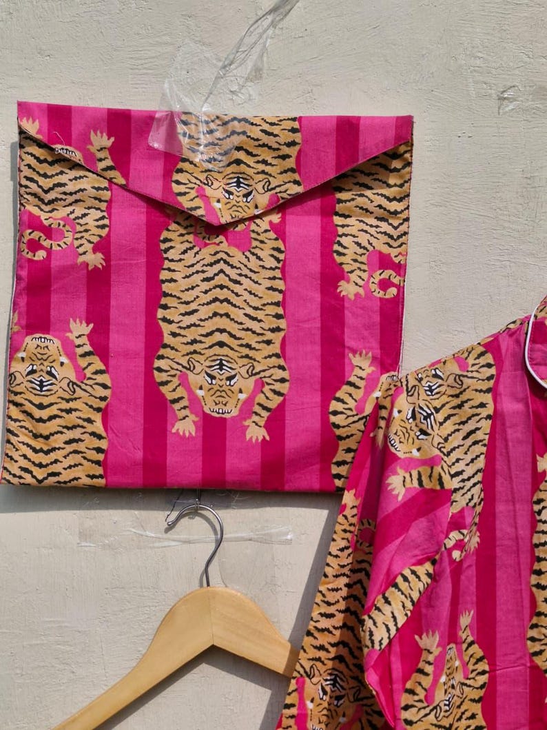 May include: Pink and brown tiger print pajama set. The set includes a button-down shirt and matching pants, with a repeating tiger pattern on a pink striped background. The set is displayed with a matching envelope.