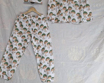 Puede incluir: Un conjunto de pijama blanco con un estampado repetido de setas verdes y marrones. El conjunto incluye una camisa de manga larga con botones, pantalones con cordón y pantalones cortos, todos colgados de perchas de madera.