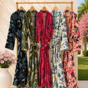 May include: Five long-sleeved robes with tie belts hanging on a wooden rack. The robes feature various colorful floral and animal prints in black, olive green, bright pink, white, and light pink, each with a contrasting striped inner lining.