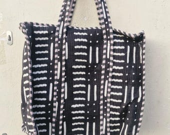 May include: A black tote bag with a white geometric pattern. The bag has two handles and a rectangular shape. The pattern consists of vertical and horizontal lines and dots. The edges of the bag and handles are trimmed with a pinkish-white fabric.