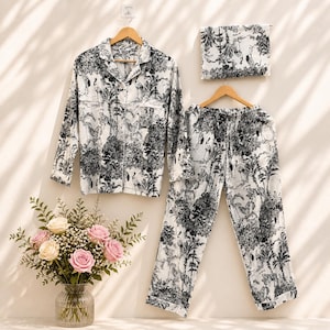 Può includere: Un completo pigiama bianco con una stampa a tema natura in bianco e nero, tra cui una camicia abbottonata a maniche lunghe, pantaloni e una mascherina per gli occhi abbinata. Il set è appeso a grucce di legno, con un bouquet di rose rosa e crema in un vaso di vetro.