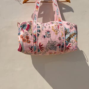 May include: A pink quilted tote bag with a floral pattern, hanging on a wooden hanger. The bag features a repeating design of flowers, birds, and other botanical elements in shades of red, blue, and green. The handles are striped.