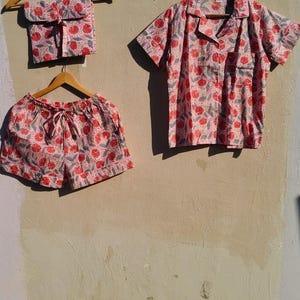 Puede incluir: Un conjunto de pijama de tres piezas con estampado floral. El conjunto incluye una camisa de manga corta con botones, pantalones cortos con cintura con cordón y una bolsa a juego. La tela es rosa claro con diseños florales rojos y grises.