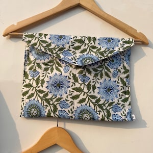 May include: A blue floral print pouch with a flap closure, hanging on a wooden hanger. The pouch features a repeating pattern of blue flowers and green leaves on a white background. The bottom of a matching garment is visible.