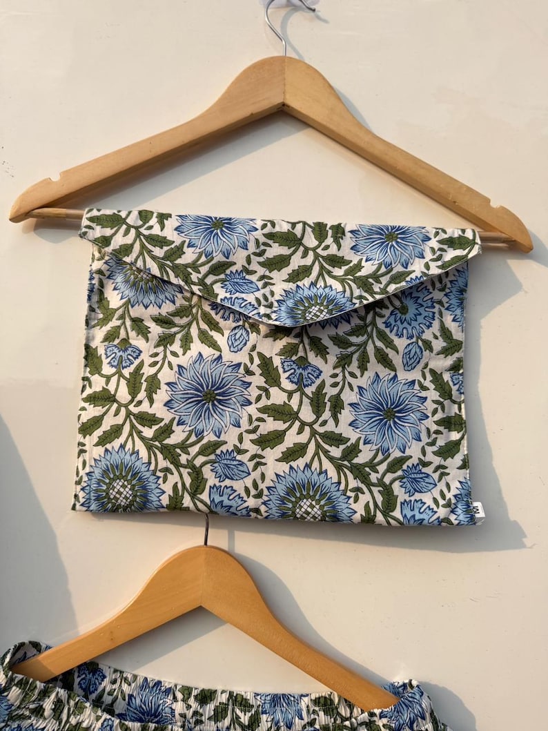 May include: A blue floral print pouch with a flap closure, hanging on a wooden hanger. The pouch features a repeating pattern of blue flowers and green leaves on a white background. The bottom of a matching garment is visible.