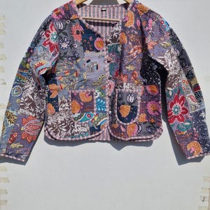 May include: A quilted jacket with a patchwork design. The jacket features a variety of floral and paisley patterns in shades of purple, blue, pink, and orange. It has a round neckline, long sleeves, and two front pockets. The jacket is hanging on a wooden hanger.