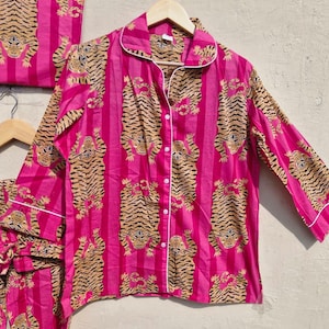 May include: A pink pajama set with a tiger print. The set includes a button-down shirt, pants, and a matching bag. The shirt and pants have a vertical stripe pattern with tiger illustrations. White trim accents the collar, cuffs, and pockets.
