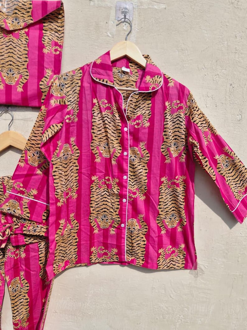May include: A pink pajama set with a tiger print. The set includes a button-down shirt, pants, and a matching bag. The shirt and pants have a vertical stripe pattern with tiger illustrations. White trim accents the collar, cuffs, and pockets.