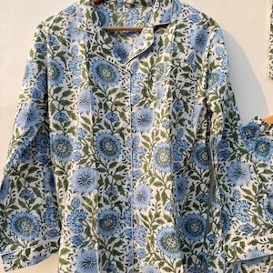 May include: A two-piece pajama set with a floral pattern. The set includes a button-down shirt and matching pants. The fabric is white with a blue and green floral design. The shirt has a collar and long sleeves. The pajamas are hanging on a wooden hanger.