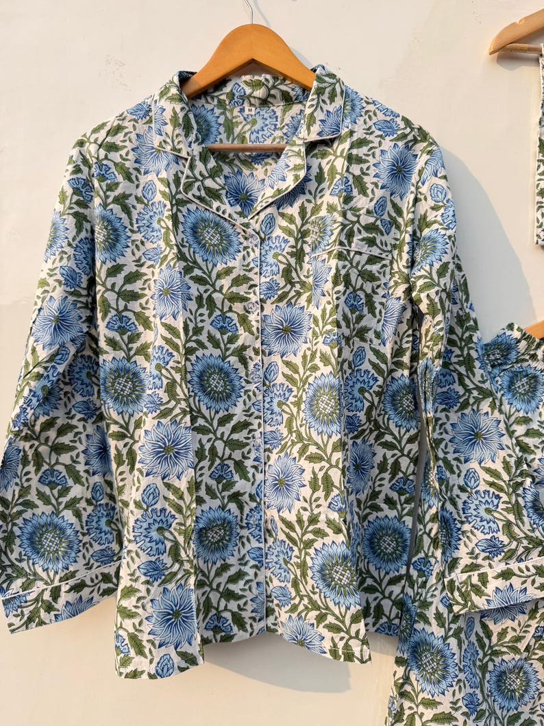 May include: A two-piece pajama set with a floral pattern. The set includes a button-down shirt and matching pants. The fabric is white with a blue and green floral design. The shirt has a collar and long sleeves. The pajamas are hanging on a wooden hanger.