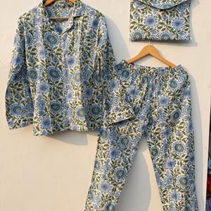 May include: A pajama set with a floral pattern in blue and green on a white background. The set includes a long-sleeved button-down shirt, pants, and a matching pouch, all hanging on wooden hangers.