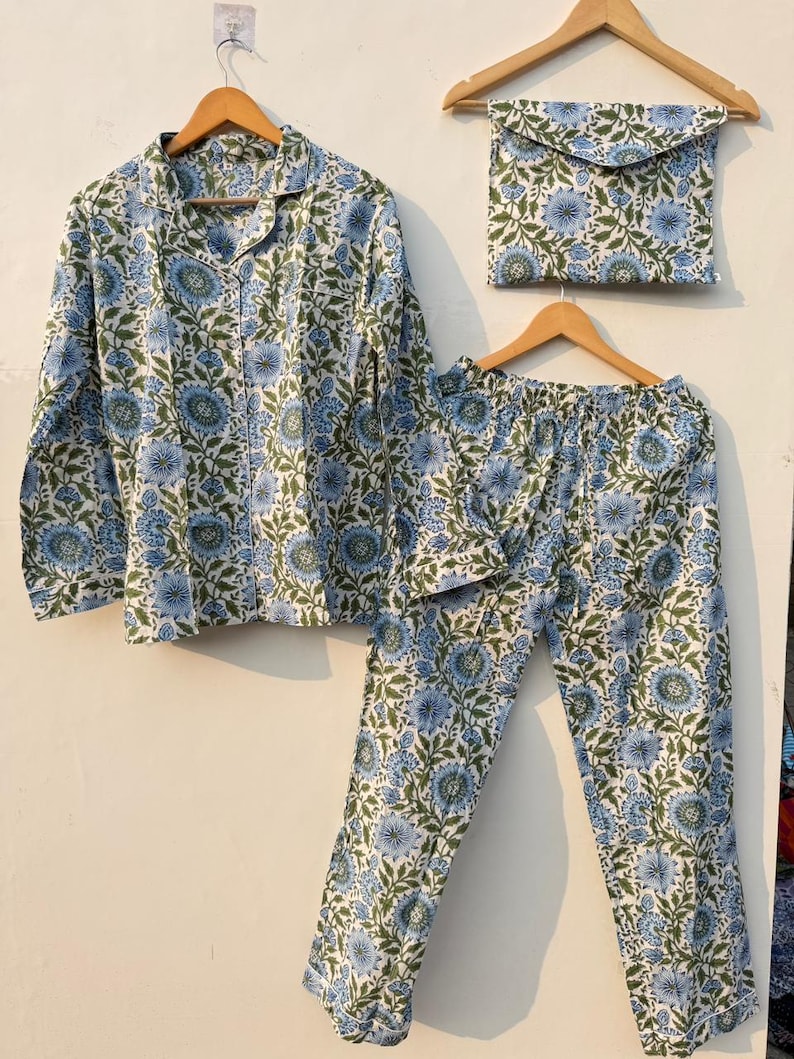 May include: A pajama set with a floral pattern in blue and green on a white background. The set includes a long-sleeved button-down shirt, pants, and a matching pouch, all hanging on wooden hangers.
