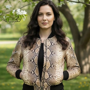 Puede incluir: Chaqueta bomber con estampado de piel de serpiente marrón y beige con ribete negro. La chaqueta tiene cierre de cremallera y puños negros. La persona lleva una camiseta negra. El fondo es una escena exterior borrosa con árboles y vegetación.