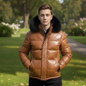 Può includere: Giacca imbottita in pelle marrone con cappuccio bordato di pelliccia nera. La giacca ha una chiusura a zip e tasche. La persona indossa una camicia nera. Lo sfondo è un parco con erba verde e alberi.