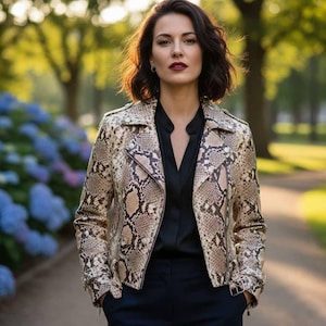 Pode incluir: Casaco de couro com padrão de pele de cobra em tons de bege e castanho, usado com uma camisa preta e calças azul-marinho. O casaco, de estilo motard, tem gola de lapela, fecho assimétrico e detalhes prateados. A mulher está num parque.