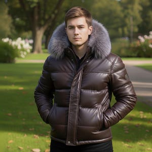 Può includere: Una giacca imbottita in pelle marrone con cappuccio foderato in pelliccia grigia. La giacca ha un design trapuntato e una chiusura frontale con cerniera. La persona è in piedi all'aperto in un parco.