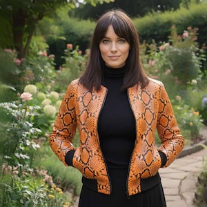 Puede incluir: Chaqueta bomber con estampado de piel de serpiente naranja y negra, con cuello y puños negros. La chaqueta está cerrada con cremallera y se lleva sobre un cuello alto negro. El fondo presenta un jardín con follaje verde y flores rosas.
