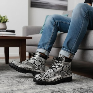 Könnte beinhalten: Kampfstiefel mit Schlangenmuster, mit schwarzen Schnürsenkeln und Sohlen. Die Stiefel haben ein schwarz-weißes Schlangenhautmuster und einen schwarzen Kragen. Die Stiefel werden mit einer blauen Jeans getragen.