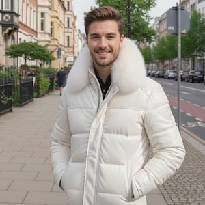 Può includere: Un piumino bianco con un ampio colletto in pelliccia bianca e soffice. La giacca ha un design trapuntato e una chiusura con cerniera frontale. La persona è in piedi su un marciapiede di fronte a una strada con edifici.