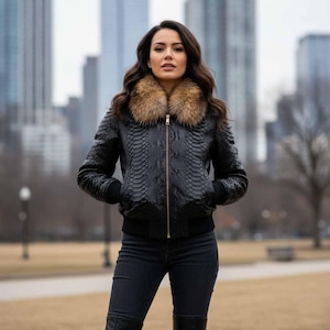 Peut inclure: Veste de bombardier en cuir noir avec un motif peau de serpent et un col en fourrure. La veste a une fermeture éclair dorée et des poignets et un ourlet côtelés noirs. La personne porte un jean et des bottes noirs.