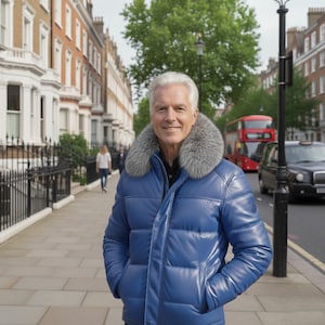 Può includere: Un uomo indossa un piumino blu con collo in pelliccia grigia, in piedi su un marciapiede di fronte a una strada con edifici e un autobus rosso a due piani. La giacca ha un design trapuntato.