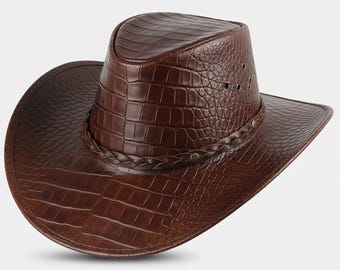 Può includere: Un cappello da cowboy in pelle marrone con un motivo strutturato a forma di coccodrillo. Il cappello presenta un'ampia tesa, una fascia intrecciata attorno alla corona e piccoli fori di ventilazione. Il cappello è di un ricco colore marrone scuro.