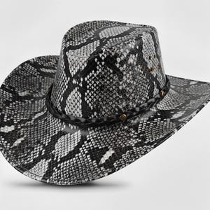Może przedstawiać: Kapelusz kowbojski z wzorem skóry węża w kolorach czarnym, białym i szarym. Kapelusz posiada pleciony pasek wokół korony i małe, złote nity. Rondo jest szerokie i zakrzywione.
