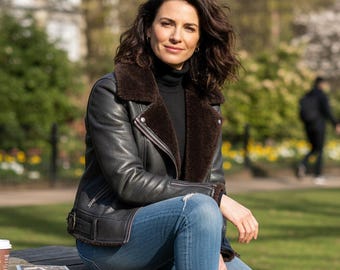 May include: A black leather jacket with a brown shearling collar and lining. The jacket has a zipper closure and is worn over a black turtleneck. The person is wearing blue jeans with distressed details. A coffee cup and a book are on the wooden bench.