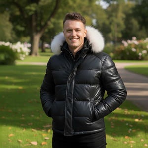 Può includere: Una giacca puffer in pelle nera con cappuccio foderato in pelliccia bianca. La giacca ha un design trapuntato e una chiusura con cerniera frontale. La persona indossa pantaloni neri. Lo sfondo è un parco con erba verde e alberi.