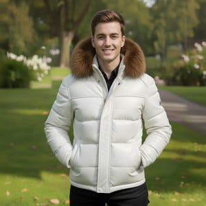 Può includere: Giacca trapuntata bianca con collo in pelliccia marrone. La giacca ha una chiusura a zip frontale e tasche. La persona indossa una camicia nera sotto. Lo sfondo è un parco con erba verde e alberi.