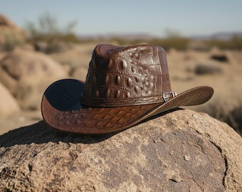 Puede incluir: Un sombrero de vaquero marrón con un patrón texturizado similar al de un cocodrilo. El sombrero presenta un ala curvada y una banda decorativa con una hebilla plateada. El sombrero descansa sobre una gran roca en un paisaje desértico.