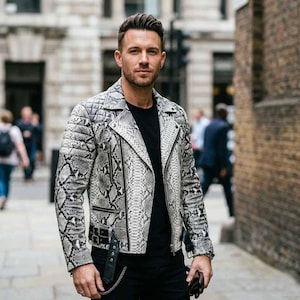 May include: A black and white snakeskin-patterned leather jacket with a quilted shoulder design. The jacket has a zipper closure, multiple pockets, and a belt detail at the waist. The person is wearing a black t-shirt and black pants.