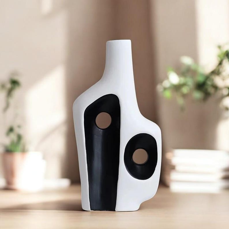 May include: A modern, abstract ceramic vase. The vase is primarily white with a bold black design featuring two circular cutouts. The vase has a narrow neck and a unique, asymmetrical shape, suitable for home decor.