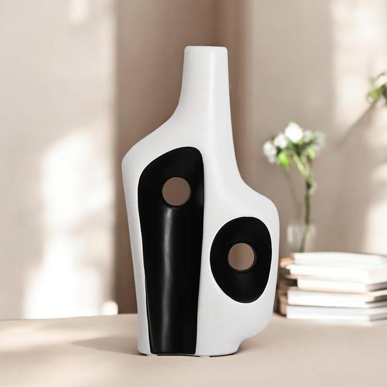 May include: A modern, abstract ceramic vase in white with black accents. The vase features a unique shape with two circular cutouts and a black vertical stripe. The vase is a decorative item for home decor.