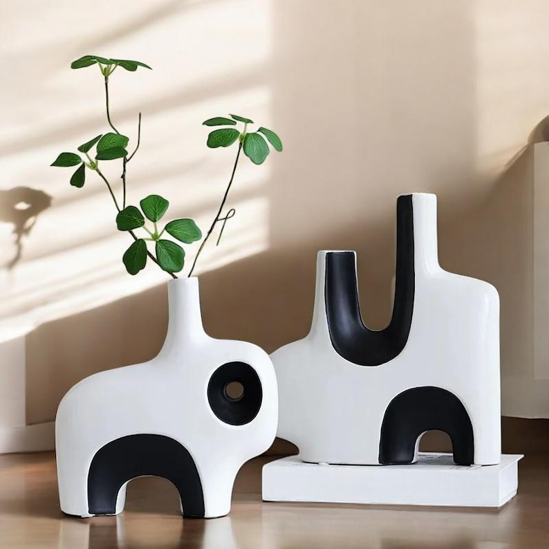 May include: Two modern white ceramic vases with black geometric accents. One vase has a circular opening and a curved base, while the other has a U-shaped design. Green leafy stems are placed in the vase with the circular opening.