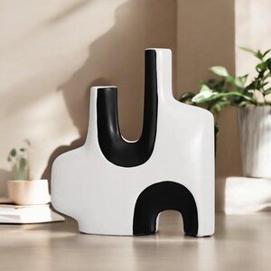 May include: A modern white ceramic vase with a bold black abstract design. The vase features a curved, organic shape with a black U-shaped accent and a black semi-circle at the base. The vase is a decorative home accent.