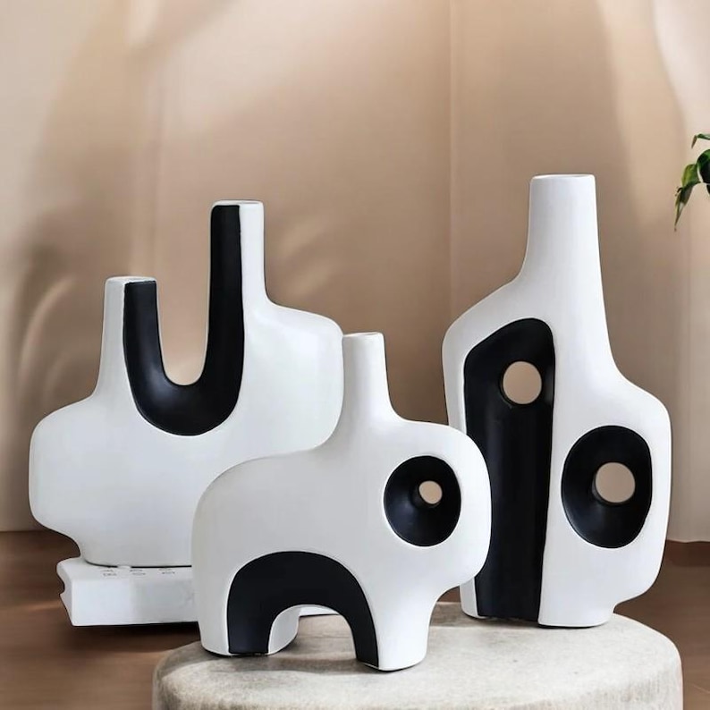 May include: Three modern ceramic vases in white with black geometric designs. The vases have unique shapes, including a U-shaped design, a rounded form with a circular opening, and a tall, abstract shape with two circular cutouts. The vases are displayed on a neutral surface.