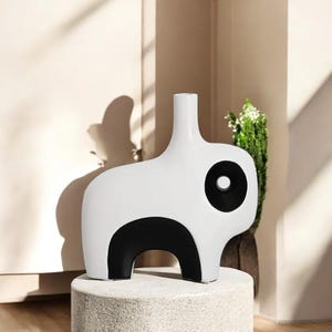 May include: A modern white ceramic vase with a black accent design. The vase has a unique shape with a rounded body and a tall, slender neck. The black accents create a striking contrast against the white background. The vase is displayed on a stone pedestal.