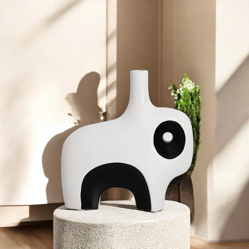 May include: A modern white ceramic vase with a black accent design. The vase has a unique shape with a rounded body and a tall, slender neck. The black accents create a striking contrast against the white background. The vase is displayed on a stone pedestal.