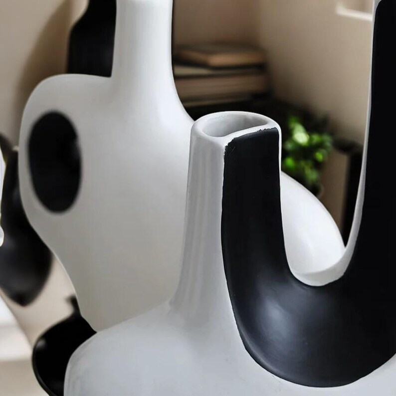 May include: A collection of modern ceramic vases in a minimalist style. The vases are primarily white with bold black accents and shapes, including a circular cutout and curved forms. The design is sleek and contemporary, suitable for home decor.