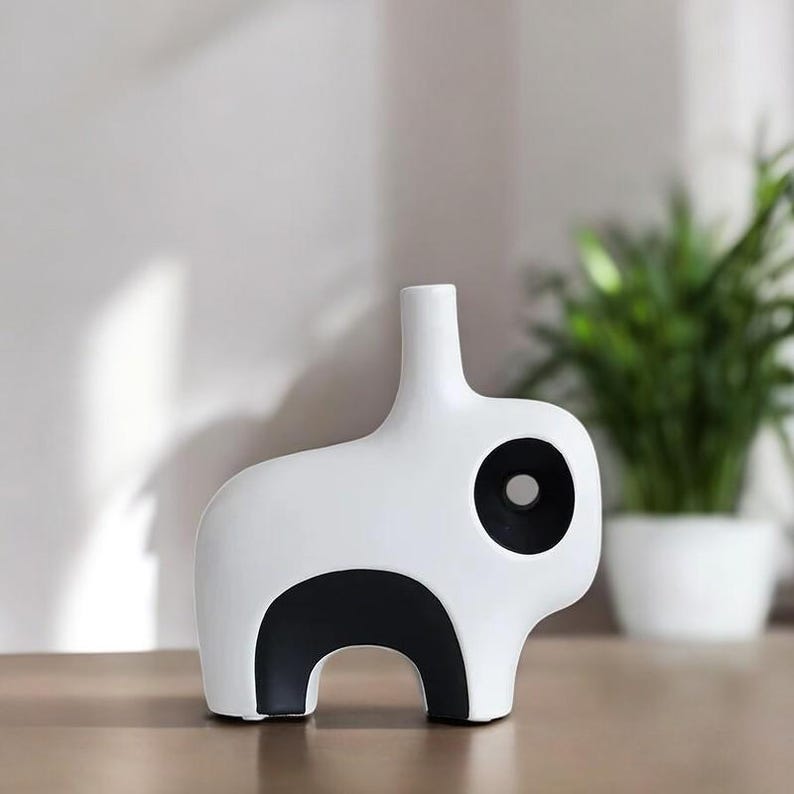 May include: A modern, white ceramic vase in the shape of an animal, featuring a black circular accent and a black curved base. The vase has a slender neck and is displayed on a wooden surface.